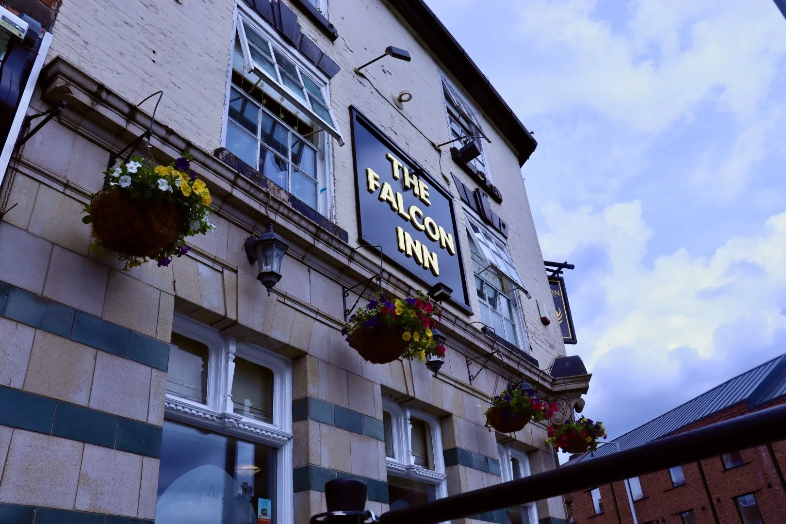 Exterior of The Falcon Inn pub showing hanging flower baskets, windows, and a black sign with gold lettering.