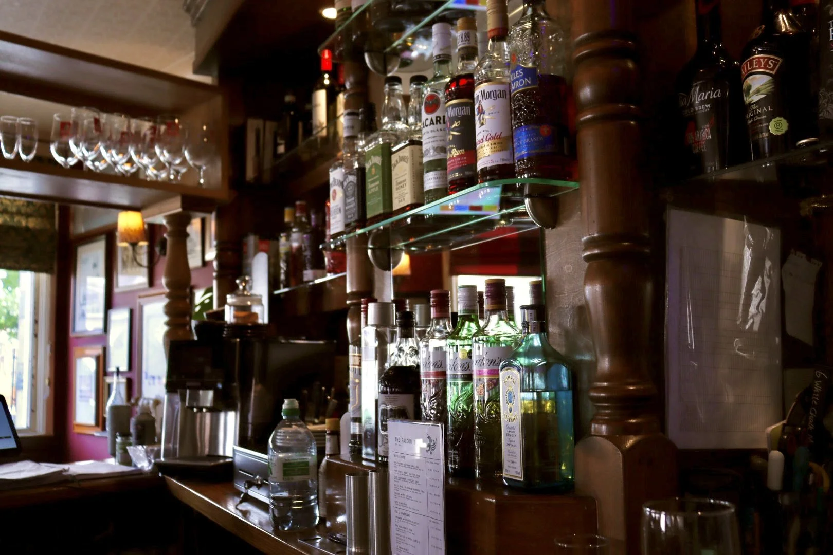 Bar area with liquor bottles on glass shelves, wine glasses hanging upside down, and various bar tools and drinks on the wooden counter, inside a cozy, dimly-lit pub or restaurant.