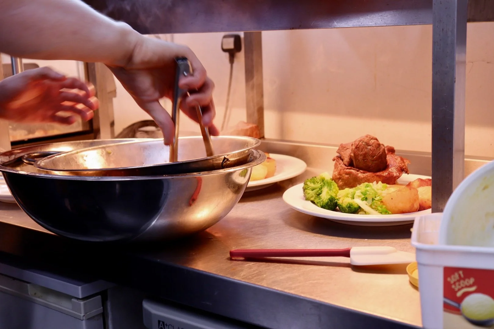 A person is serving food from a large metal bowl onto a plate. On the counter, there is a plate with broccoli, potatoes, and a piece of roast beef.