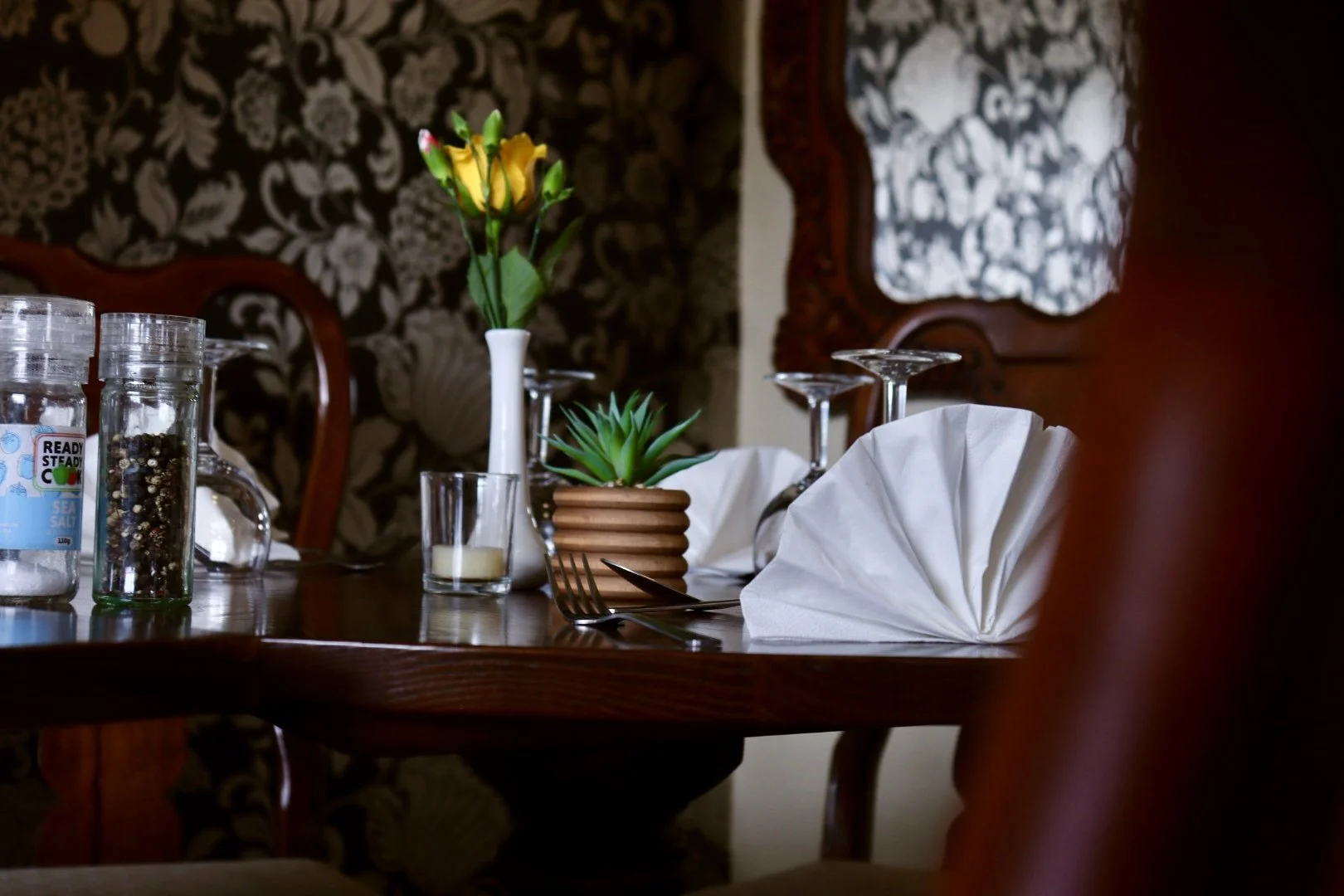 A dining table set with a yellow flower, a small green succulent, wine glasses, a fork, and a folded white napkin. In the background are patterned curtains and wooden furniture.