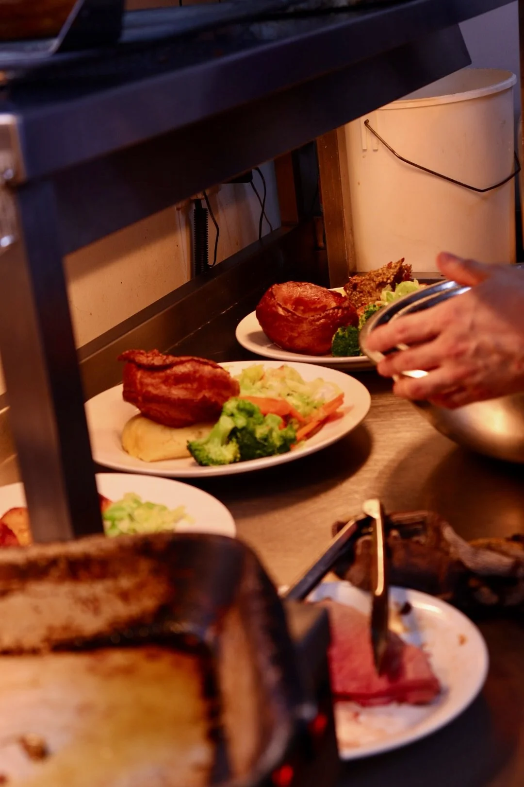 Serving station with plates of cooked roast beef, broccoli, carrots, and other vegetables, with a hand holding a metal bowl in the foreground.