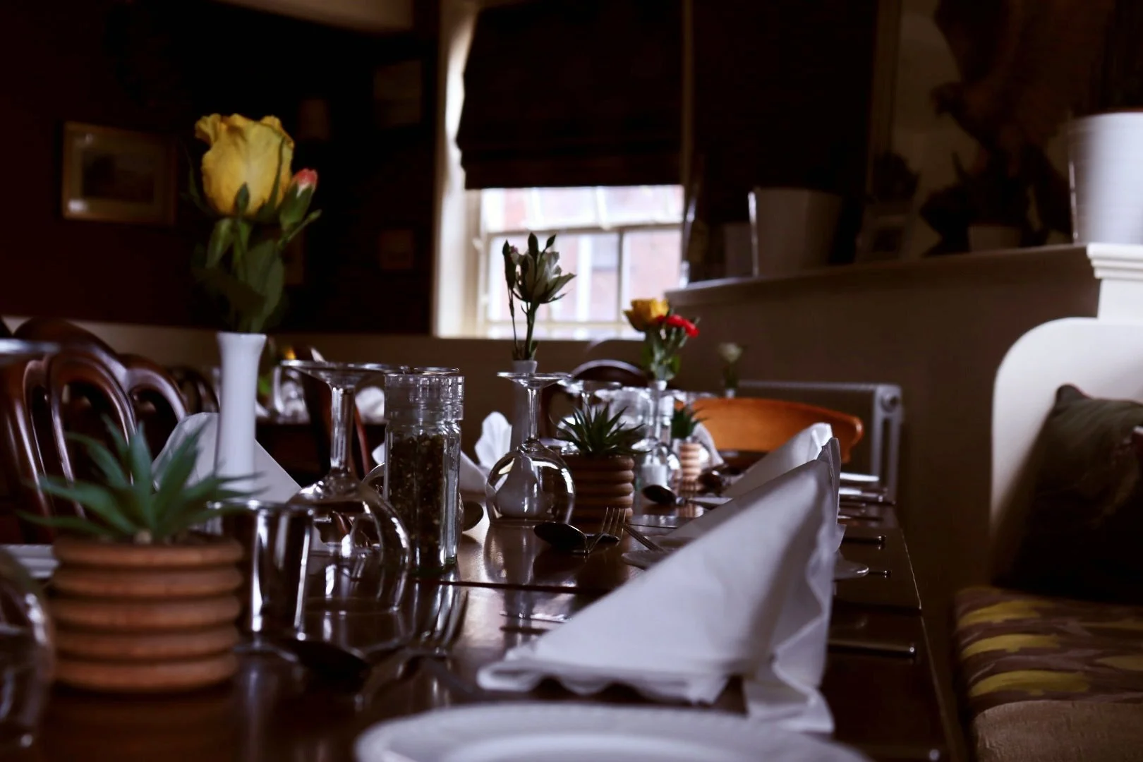 Dining table with flowers, glassware, napkins, and potted plants in a warmly lit room with window in the background.