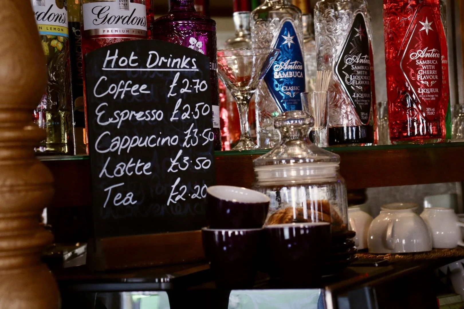 A chalkboard menu listing hot drinks, including coffees and teas with prices, placed in front of a bar shelf containing bottles of alcohol, cocktail glasses, white cups, and a jar with biscuits.