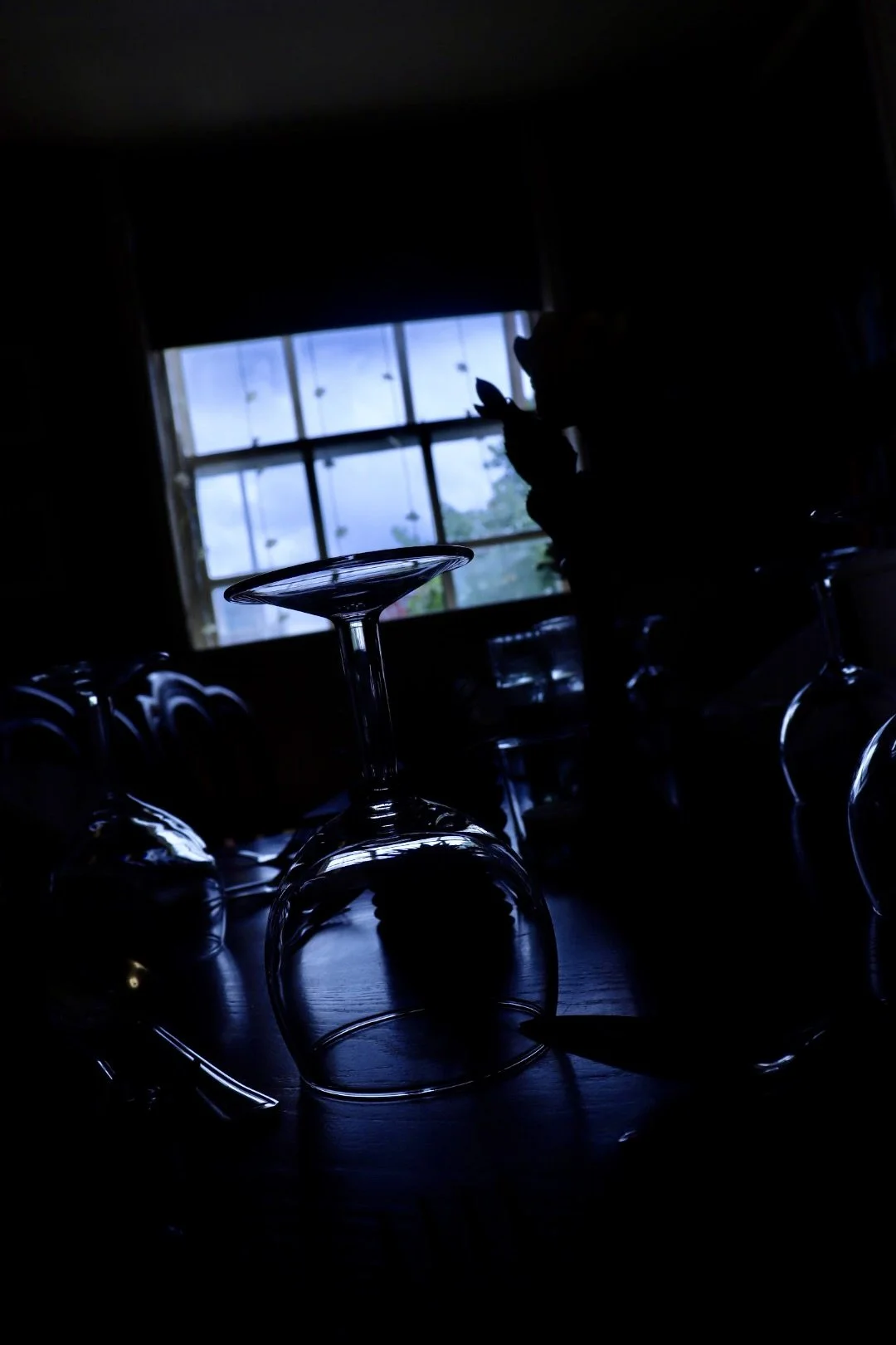 Silhouette of a person looking at their phone near a window, with wine glasses on the table in front, in a dimly lit room.