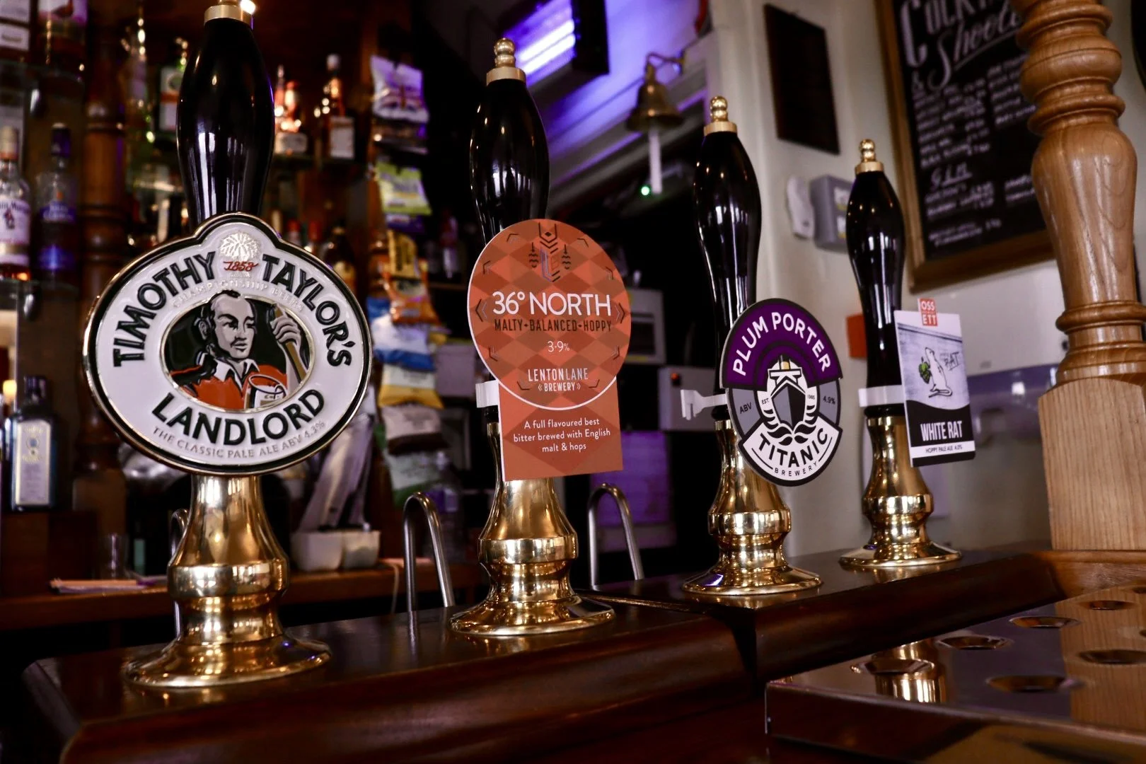 Four beer taps with labels for Timothy Taylor's Landlord, 36° North Malty Balanced Hoppy, Titanic Plum Porter, and White Rat. The bar area with bottles and a chalkboard menu in the background.