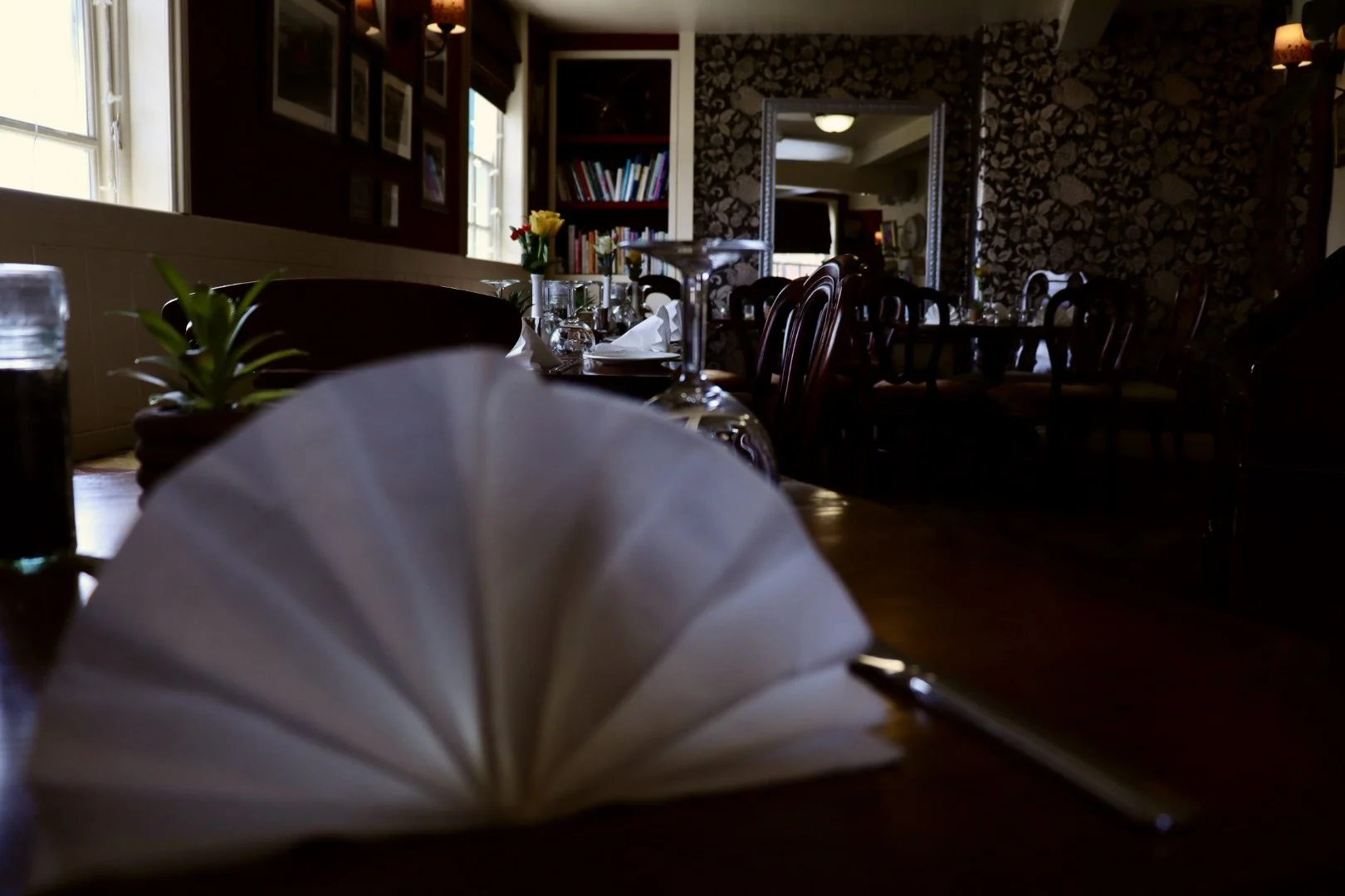 A dimly lit restaurant with a long table set for dining, decorated with flowers, tableware, and glasses. A large mirror and bookshelves are visible on the walls, and windows allow some natural light to enter.