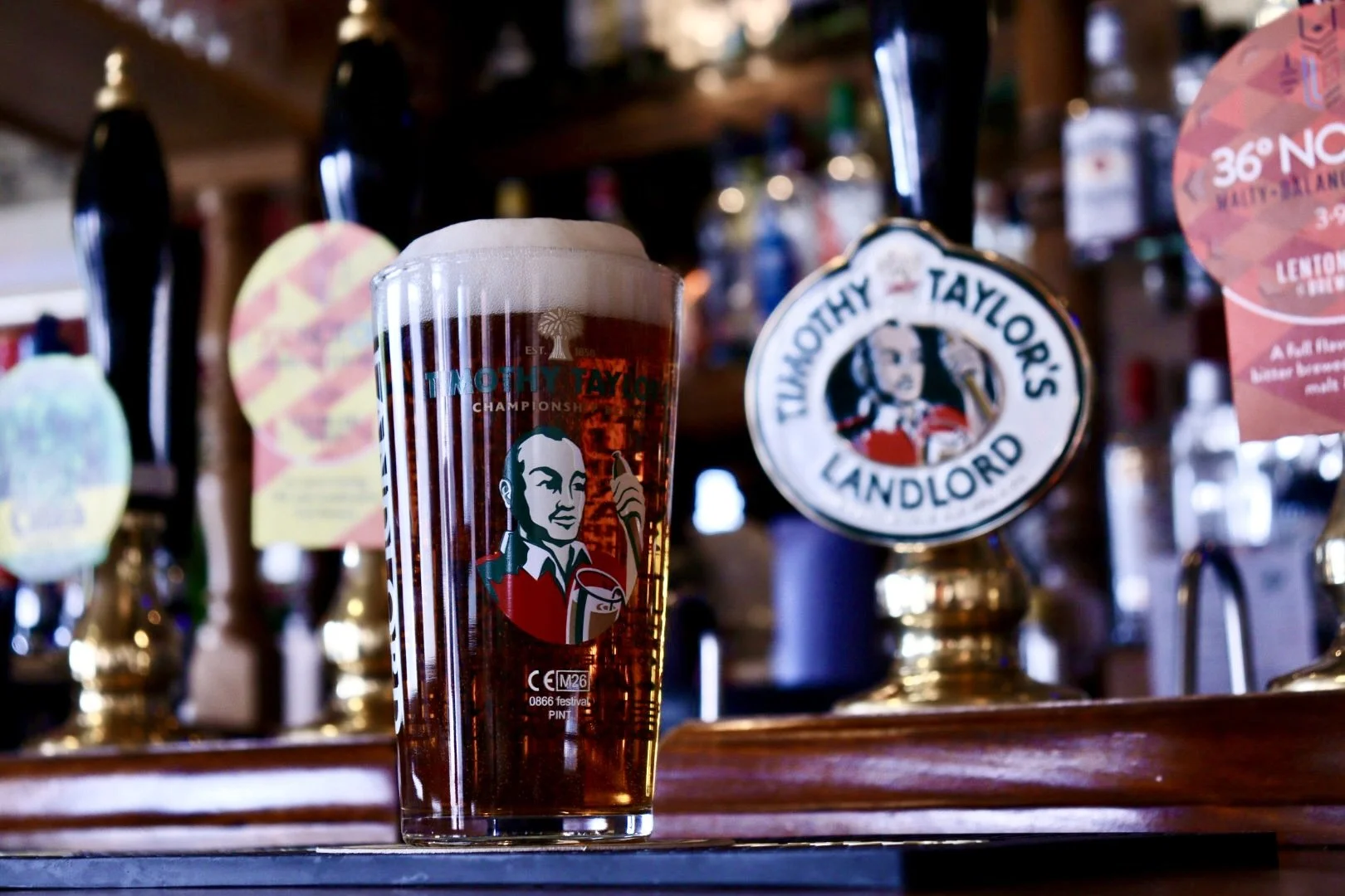 A pint glass of amber beer with foam on top, sitting on a bar counter. The glass features Timothy Taylor's logo of a woman holding a beer. In the background, there are various beer taps and bottles.
