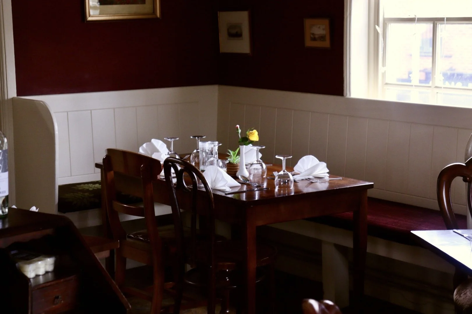 A small dining table set with white folded napkins, empty wine glasses, and a yellow flower in a white vase, inside a cozy restaurant with dark red walls and a window letting in natural light.