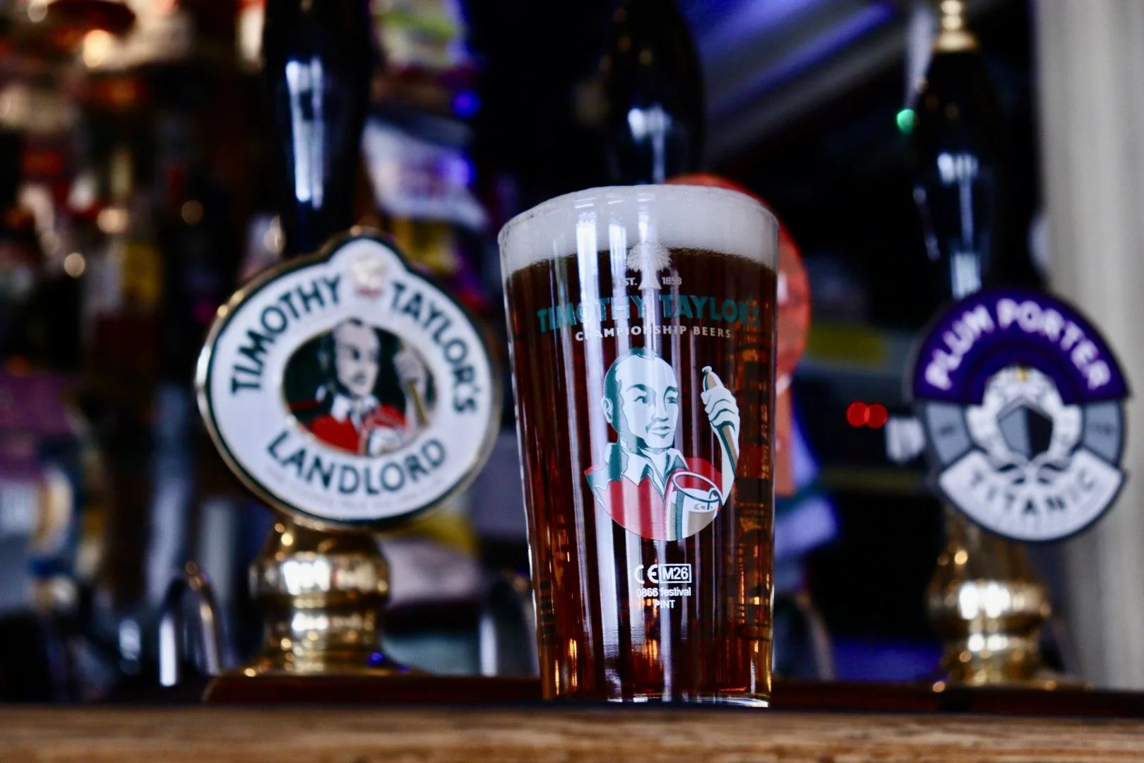 A pint glass filled with dark beer features the Timothy Taylor's logo and illustration of a woman holding beer. The background shows beer taps with labels, including Timothy Taylor's Landlord and Plum Porter.