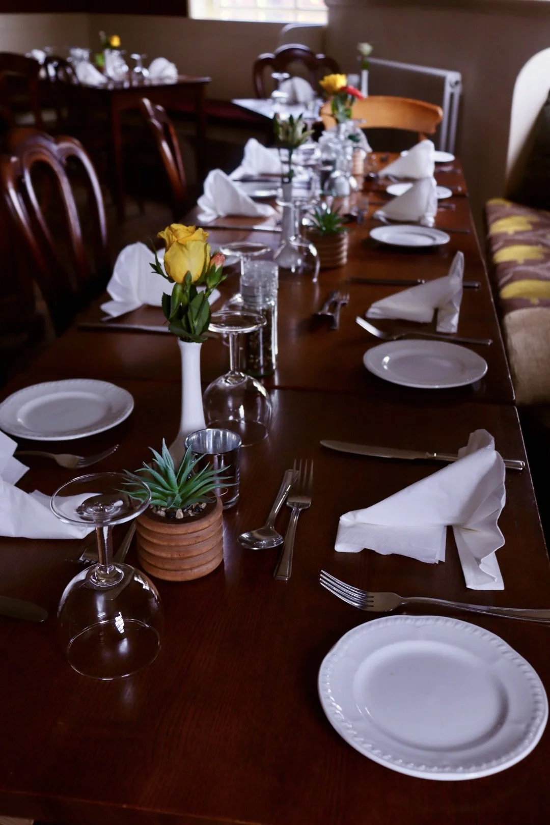 A long dining table set for a meal with white plates, silverware, folded white napkins, glasses, and small vases with yellow and white flowers; a potted succulent plant is in the foreground.