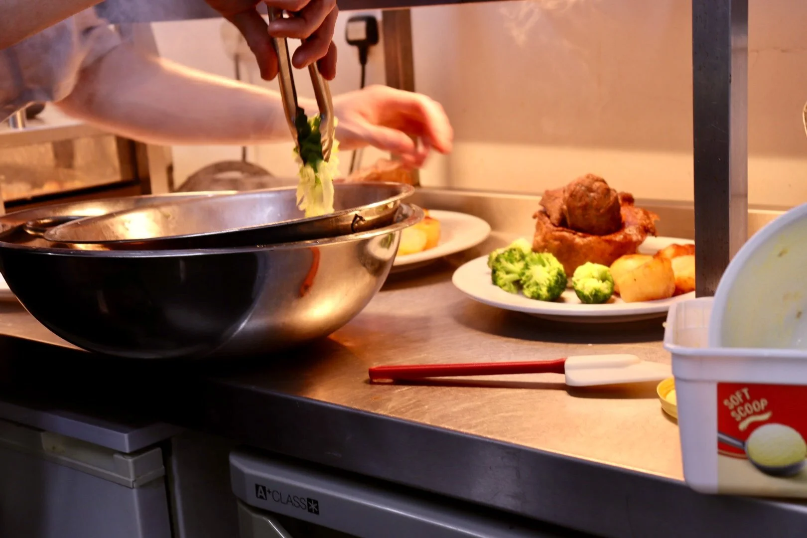 A person is serving food from a large metal bowl onto a plate. On the counter, there is a plate with broccoli, potatoes, and a piece of roast beef.