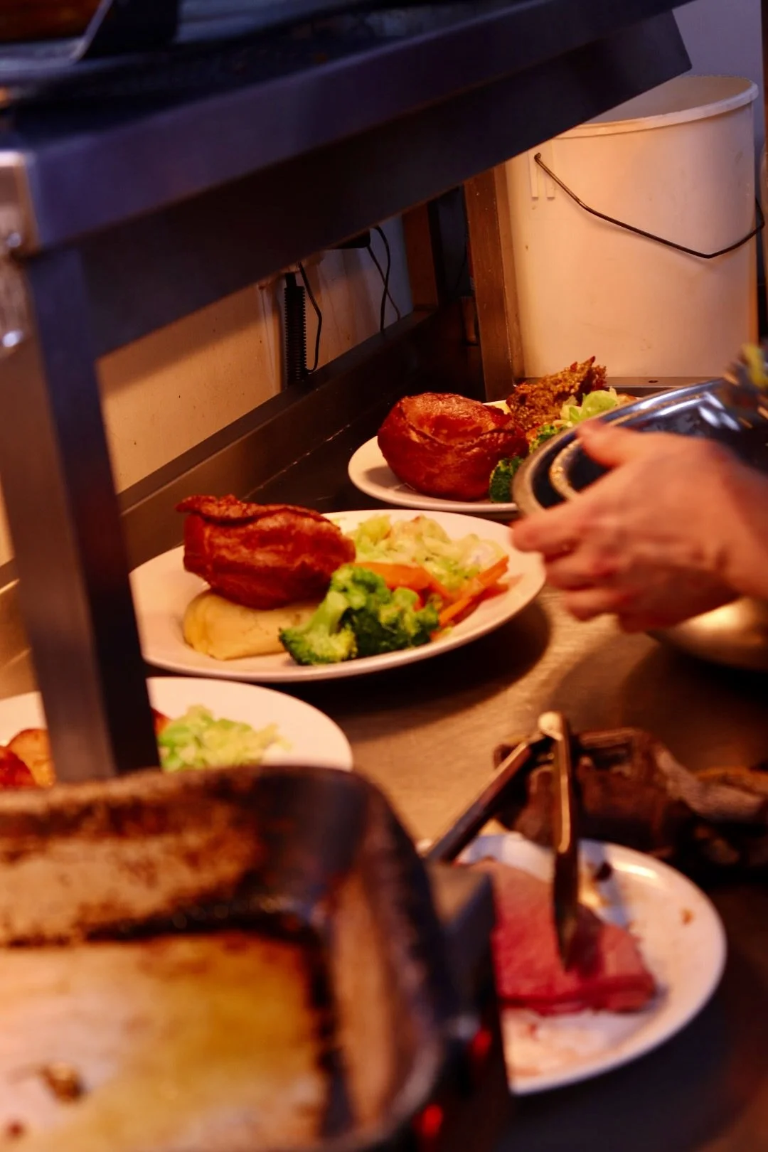 Plates of food with roast chicken, vegetables, and baked potato on a buffet line, with a hand serving more food.