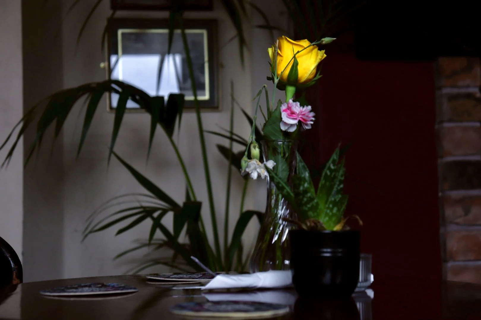Vase with yellow rose and pink flowers on a table, with a plant and window in the background.
