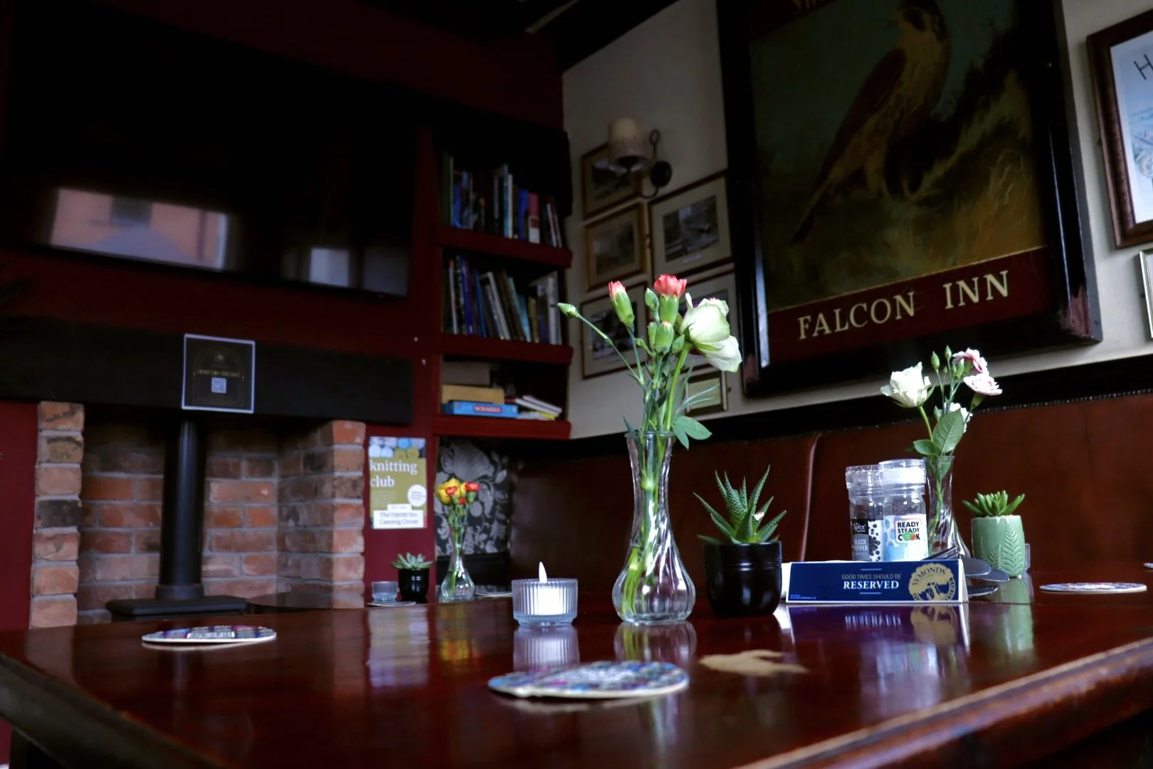 A wooden table decorated with flowers in vases, small potted plants, candles, and reserved signs inside a cosy room with framed art and a bookshelf.