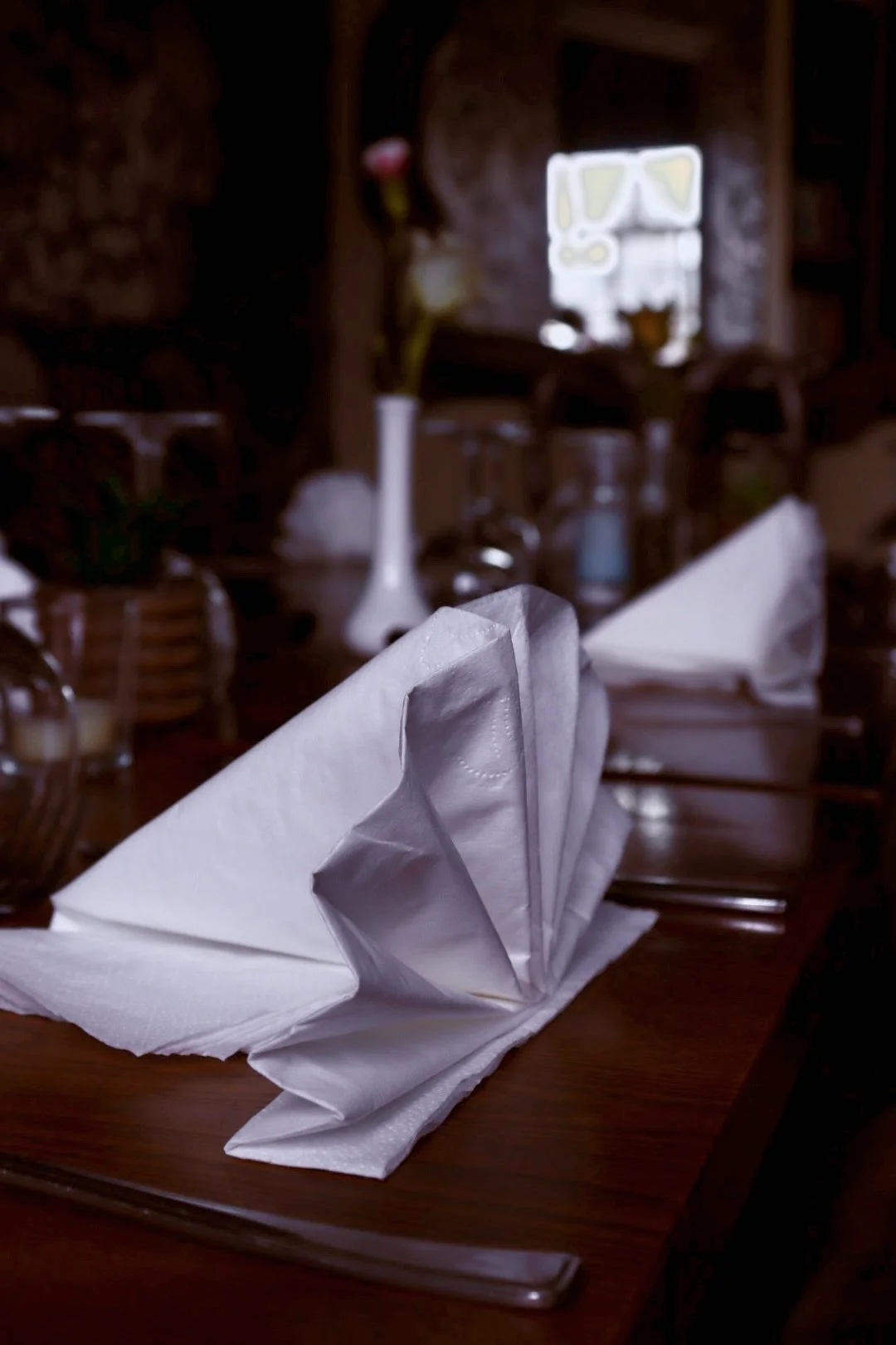 A table setting with a white folded napkin, a wooden table, and blurred decorative items and a window in the background in a cosy restaurant.