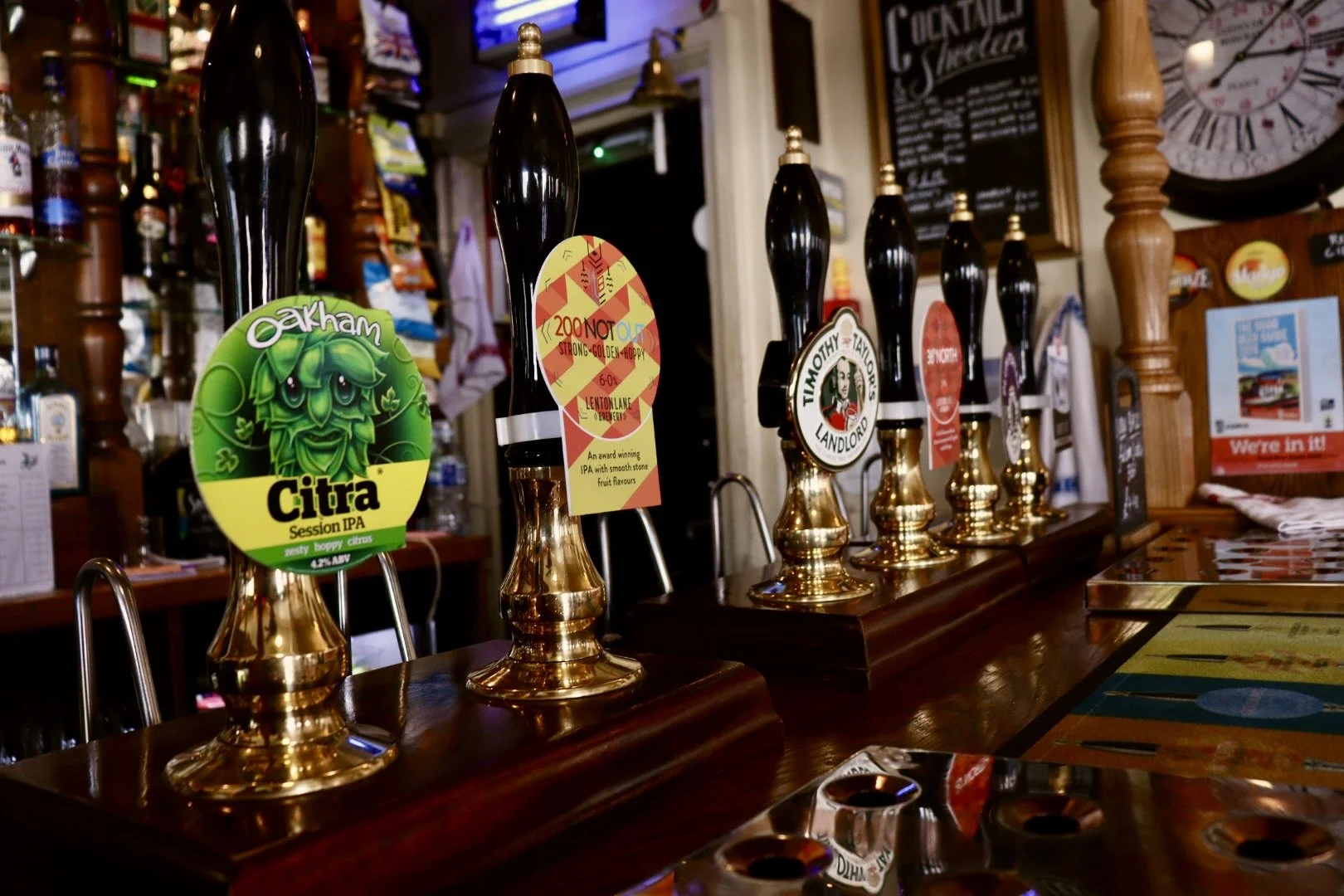 Bar countertop with several beer tap handles, each with colorful labels for different beers, in a dimly lit pub setting.