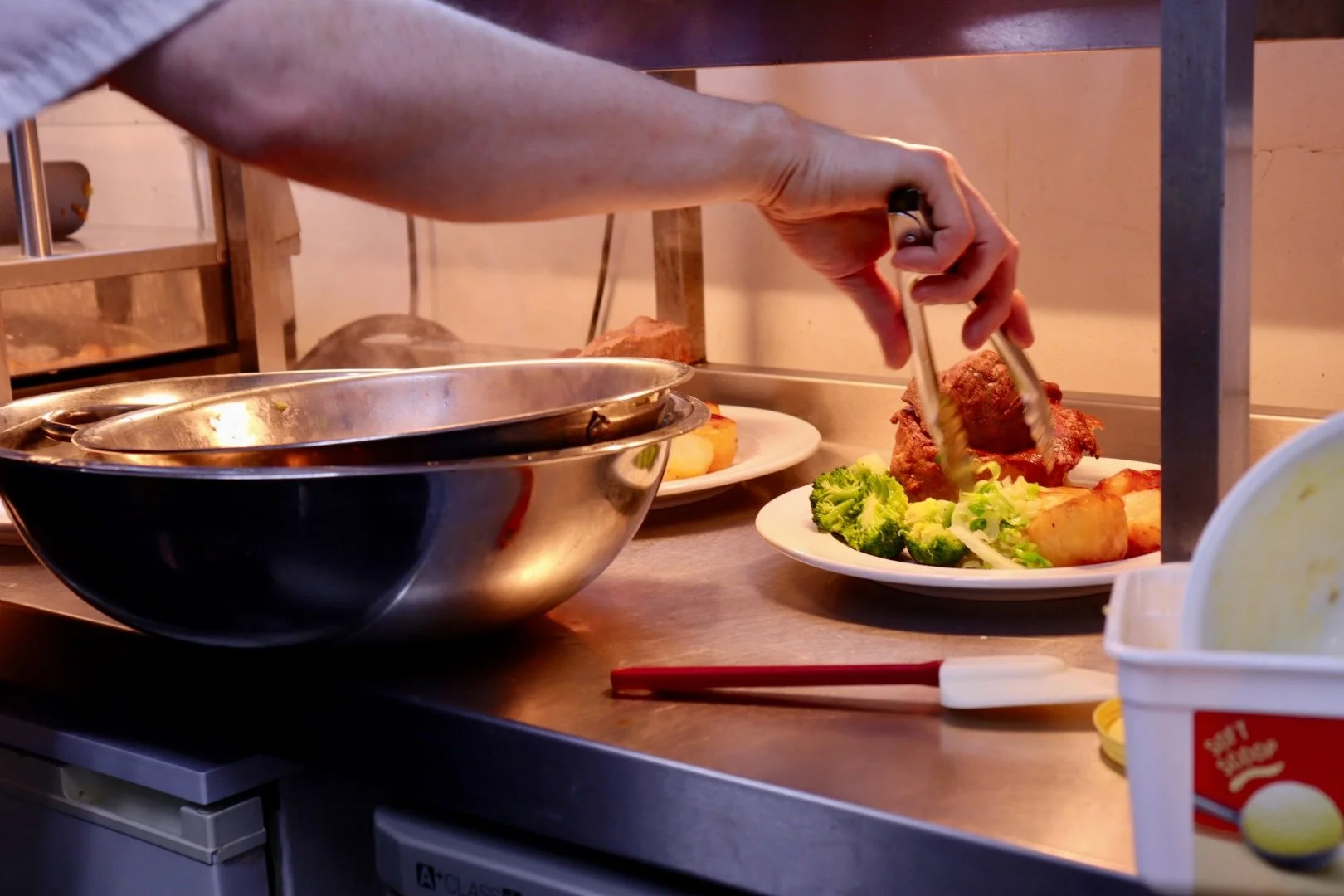 A person is serving food from a large metal bowl onto a plate. On the counter, there is a plate with broccoli, potatoes, and a piece of roast beef.