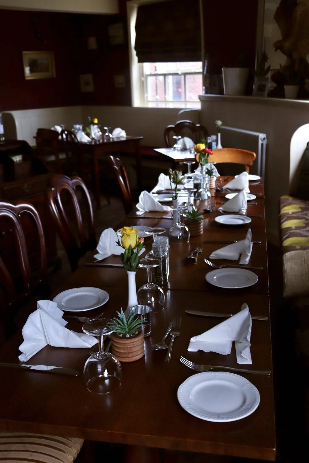 Set tables in a cosy restaurant with white napkins folded onto plates, glasses upside down, silverware arranged on each side, and small vases with colorful flowers as centerpieces, with warm wood furnishings and decor.