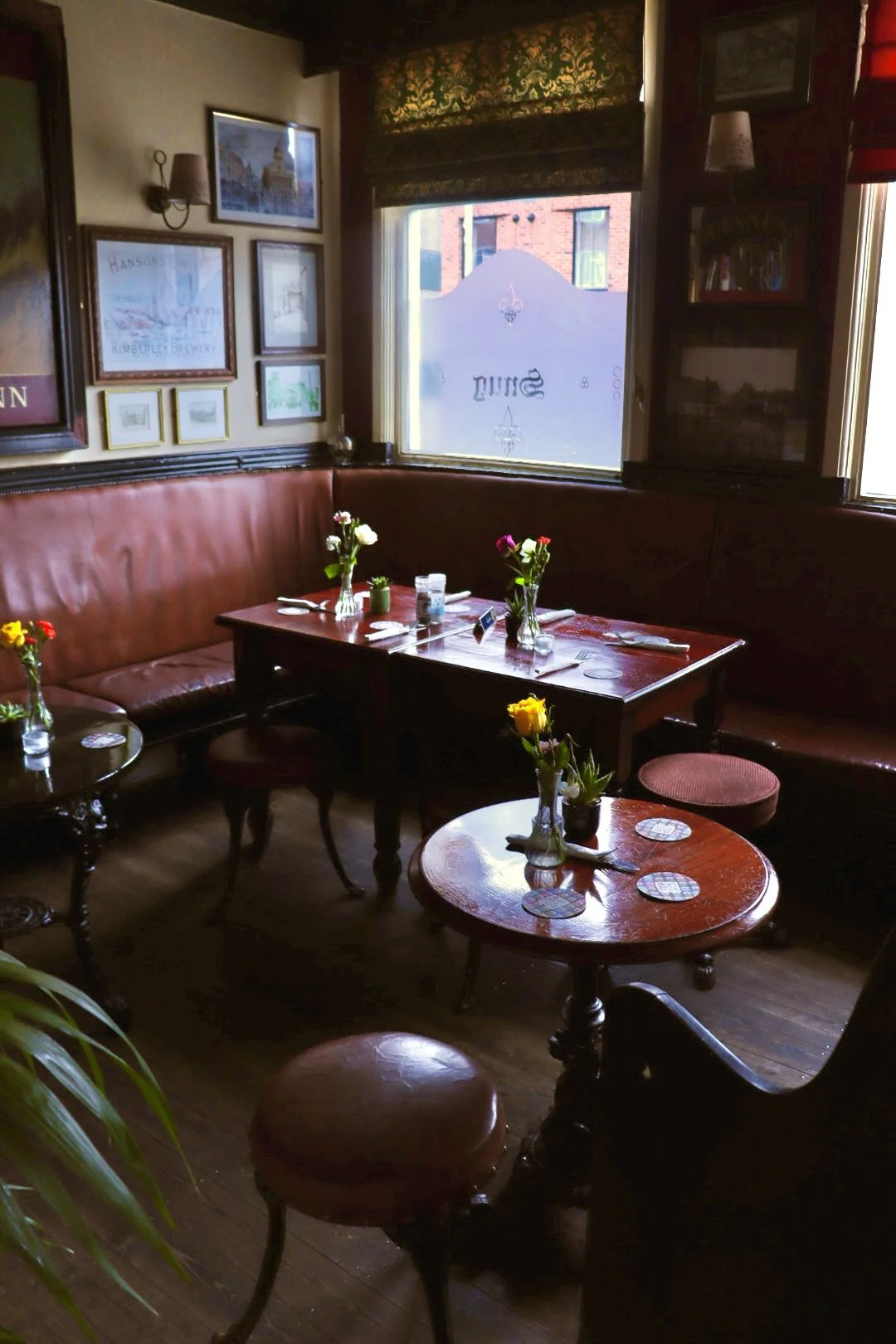 Cosy restaurant corner with wooden tables and red leather banquettes, decorated with vases of colorful flowers. Framed artwork hangs on the wall, and sunlight filters through a window with patterned curtain, illuminating the intimate dining space.