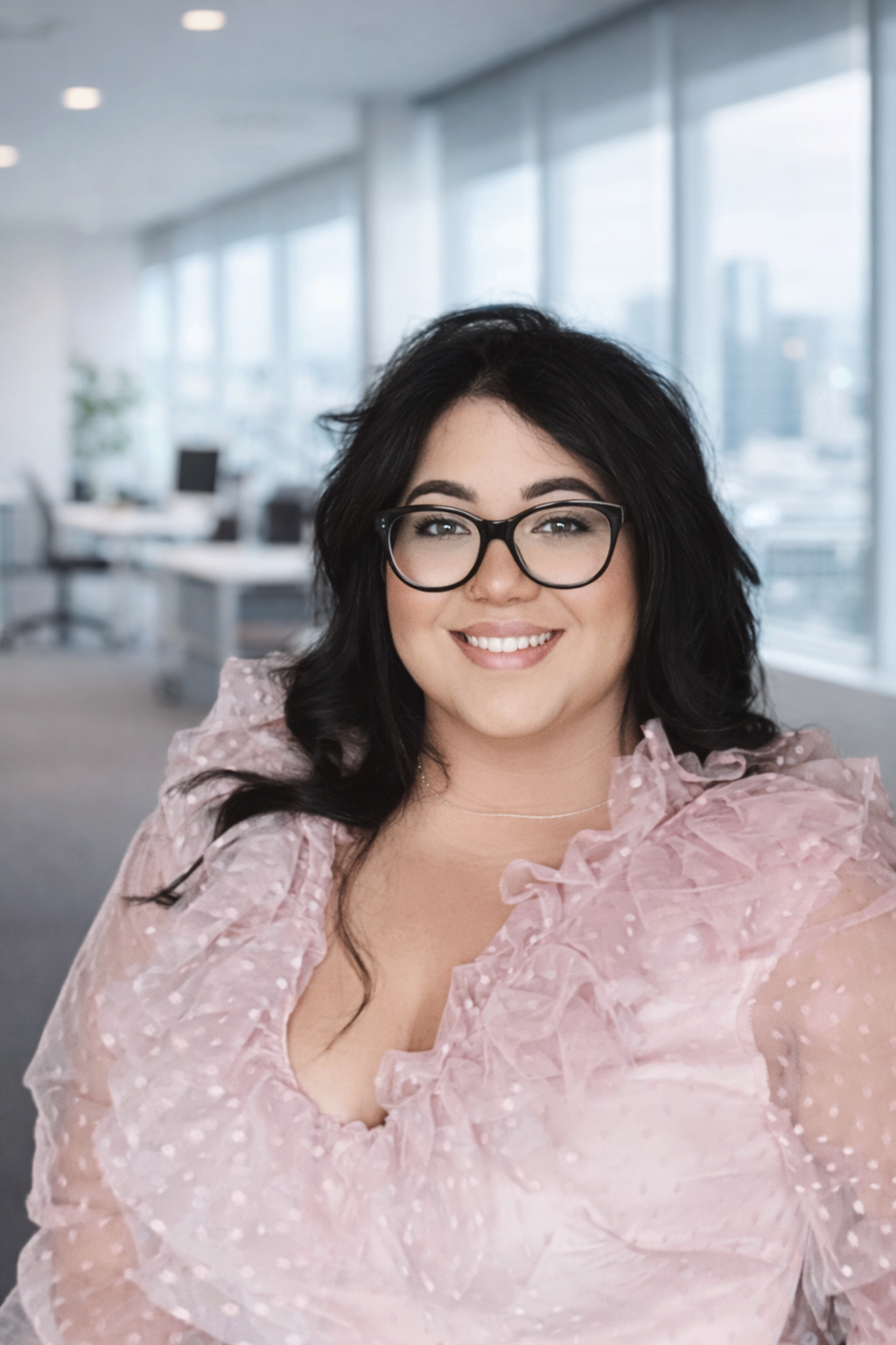 A woman with black hair, glasses, and a pink ruffled blouse smiling in an office with large windows and a city view.