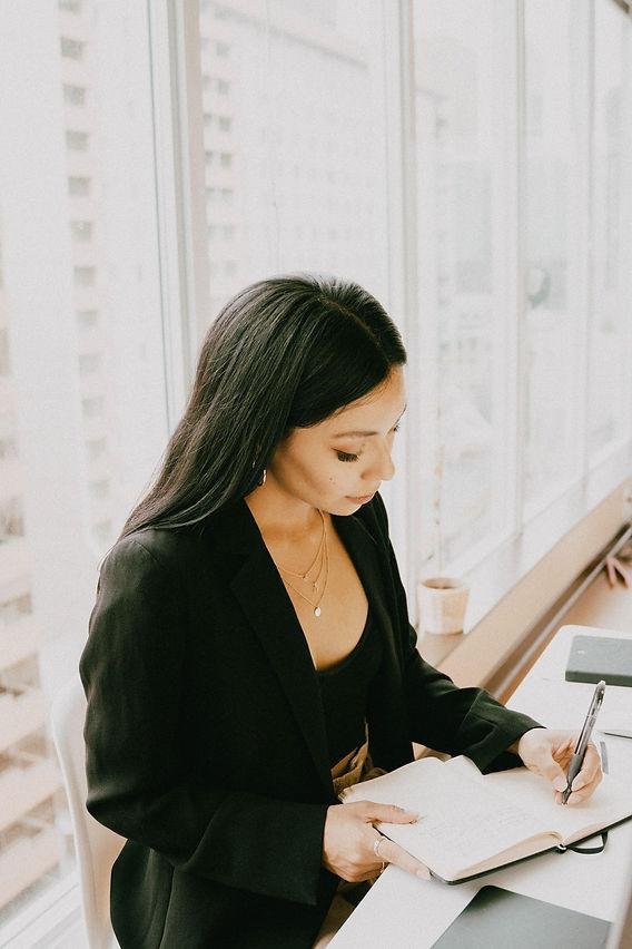A woman with long dark hair, dressed in black, sitting at a desk next to a large window, looking down at a notebook and writing with a pen.