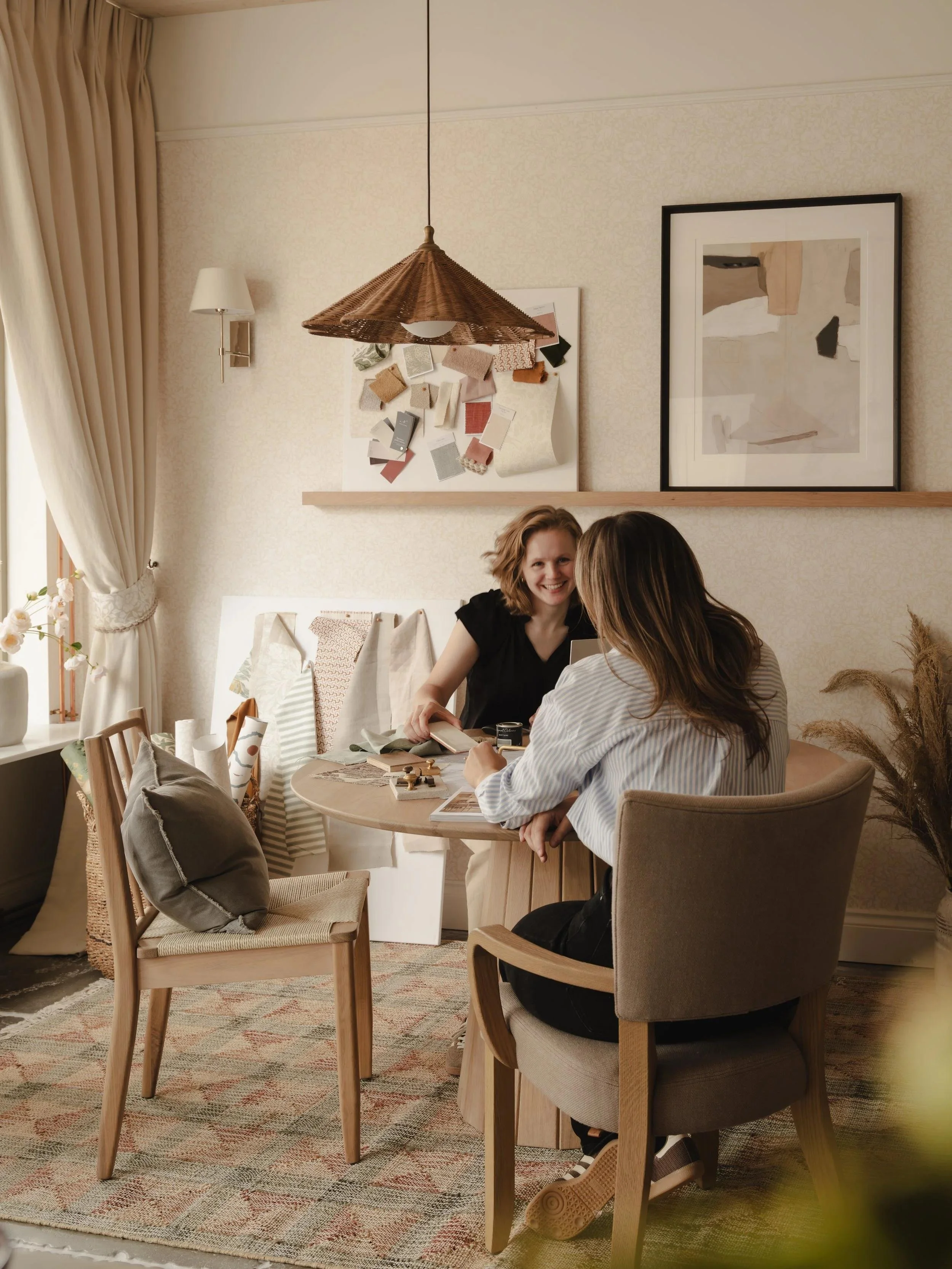 Two women sitting at a round table and talking inside a cozy, well-lit room with framed art and fabric samples on the wall.
