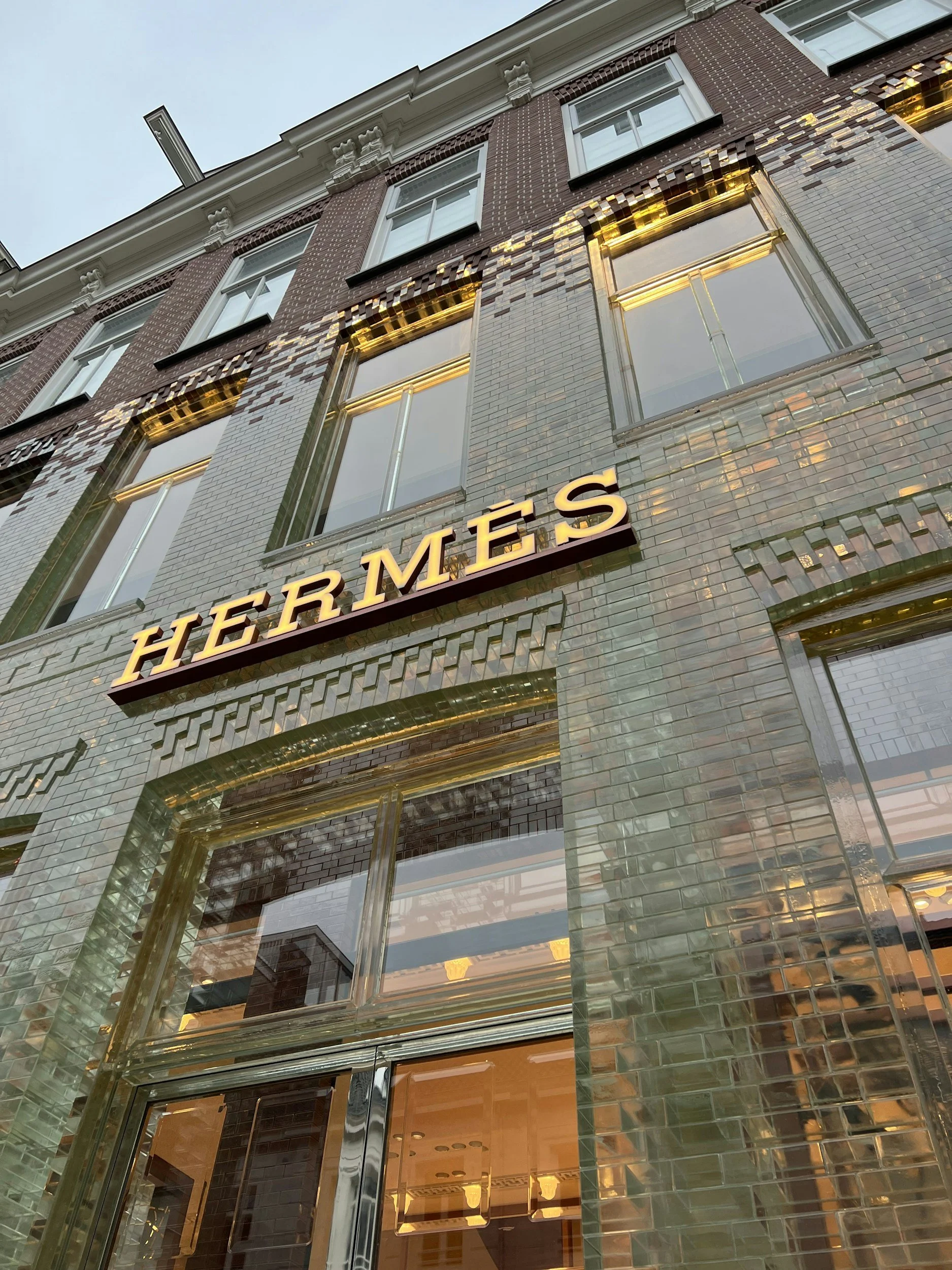 The image shows the exterior of a modern building with brick and tile facade, large glass windows, and illuminated lettering that spells out 'HERMÈS'.