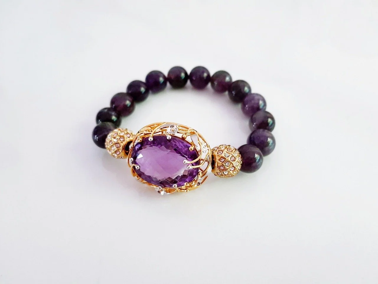 A beaded bracelet with purple beads and a large purple gemstone centerpiece set in a gold setting, accented with small clear stones.