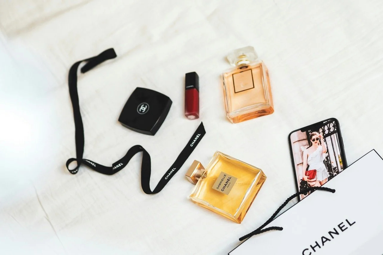 Collection of Chanel beauty and fragrance items, a lipstick, a perfume bottle, a compact, a shopping bag, a smartphone with a woman in sunglasses on the screen, and black Chanel ribbon.