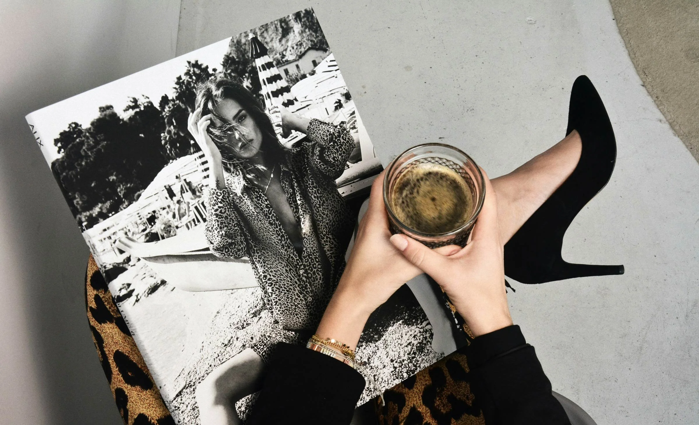 Person sitting at a table holding a glass of drink with a black and white magazine or poster featuring a woman in sunglasses and leopard print clothing. The person's legs, one in a black high heel shoe, are visible, and the floor is concrete.