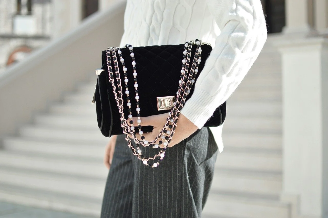 Person holding a black quilted handbag with silver hardware and layered pearl and chain necklaces, wearing a white cable-knit sweater and gray pinstripe pants.