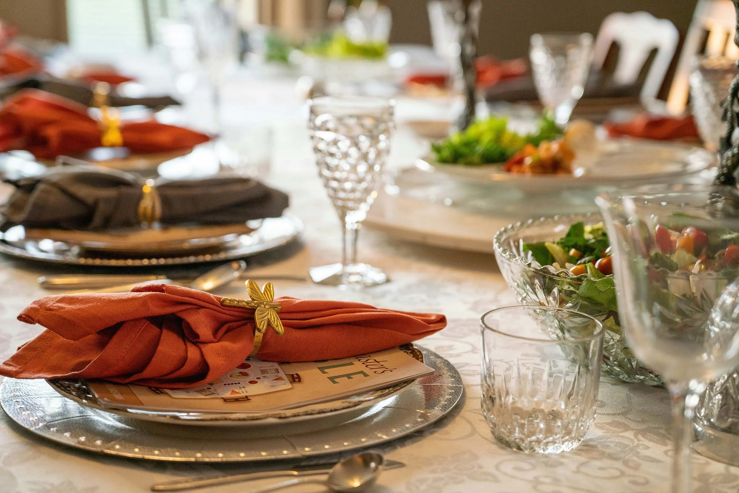 A table set with dinnerware, including a stack of plates with gold-accented black napkins and orange cloth napkins tied with gold butterfly napkin rings. There are also glassware and a large crystal bowl filled with a colorful salad. A glass of water is in the foreground, and the background shows additional table settings and greenery.