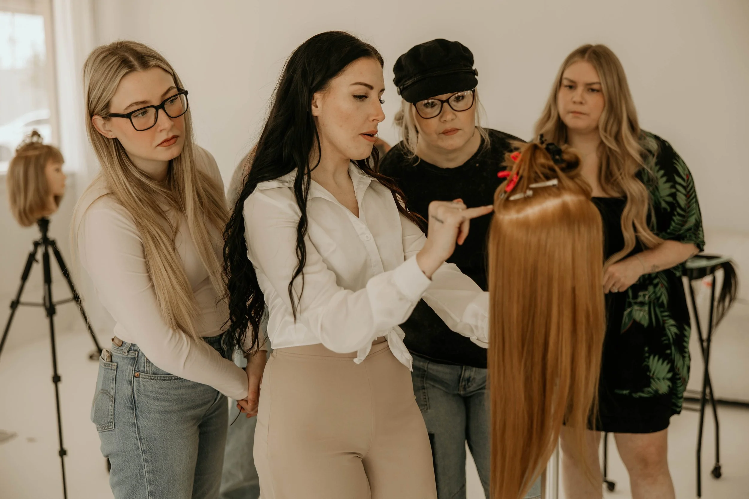 Four women appear to be having a hair consultation, focusing on a mannequin head for education on hair extensions and methods of installment