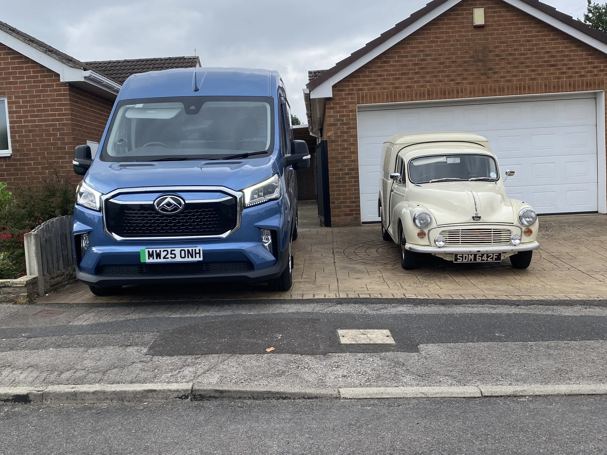 A blue delivery van and a small white vintage car parked in front of a red brick house with a white garage door.