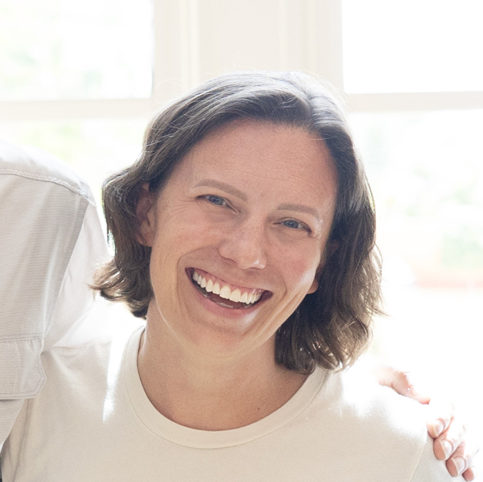 Close-up of a smiling woman with shoulder-length brown hair wearing a white shirt, in a bright room with a window in the background.