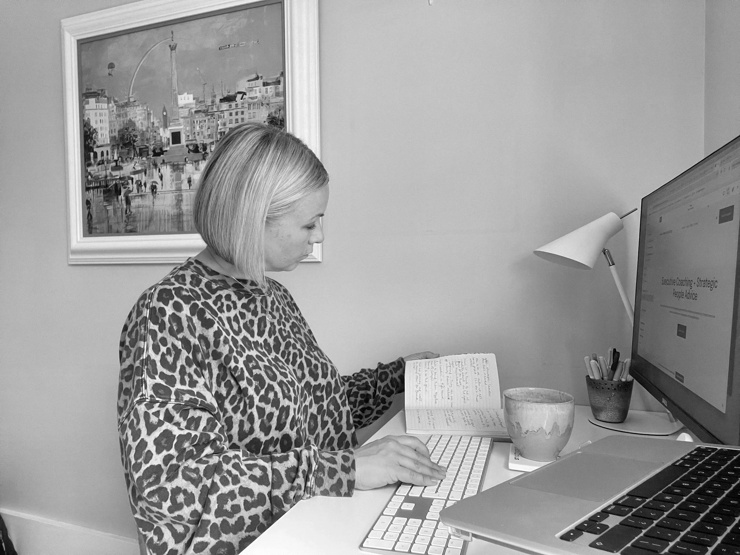 A woman with short blonde hair wearing a leopard print top, sitting at a desk, looking at a computer screen, with a notebook, two pottery mugs, and pens on the desk, in a home office with a framed cityscape painting on the wall.