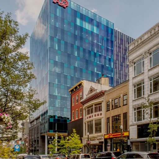 A city street with a modern glass skyscraper and older brick and white buildings, with trees and parked cars.