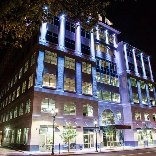 Nighttime view of a large multi-story building with illuminated blue and white lighting, large windows, and street lamps on the sidewalk in front.