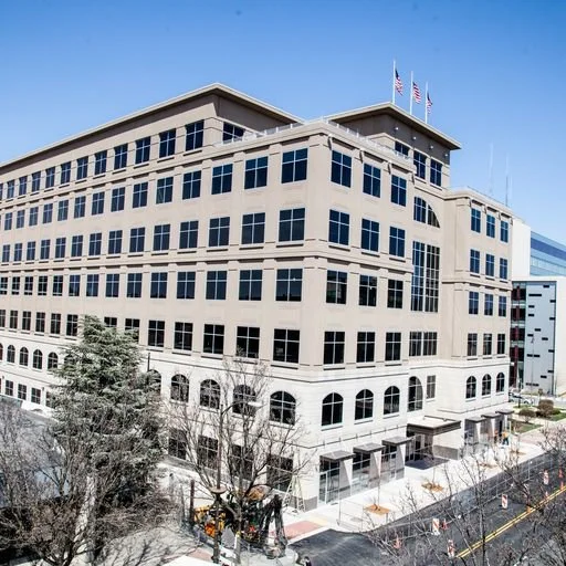 A large, multi-story modern office building with numerous windows, flags at the top, and a sidewalk in front, during the daytime with clear skies.