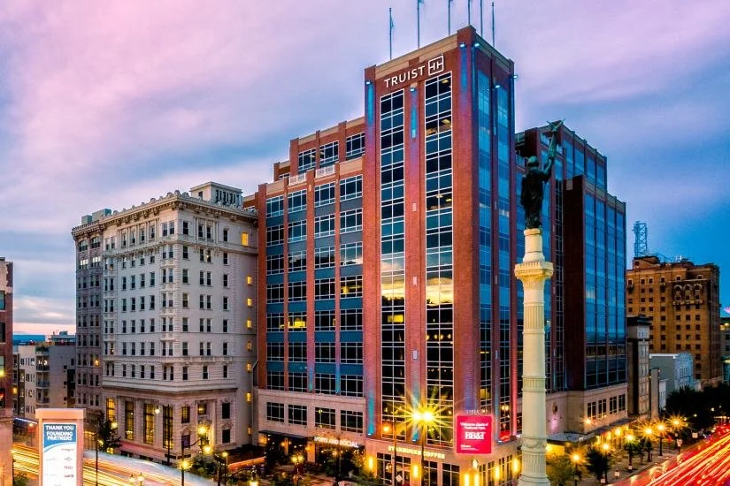 Cityscape of downtown with tall glass and brick buildings, a monument with a statue, and busy streetlights at dusk.