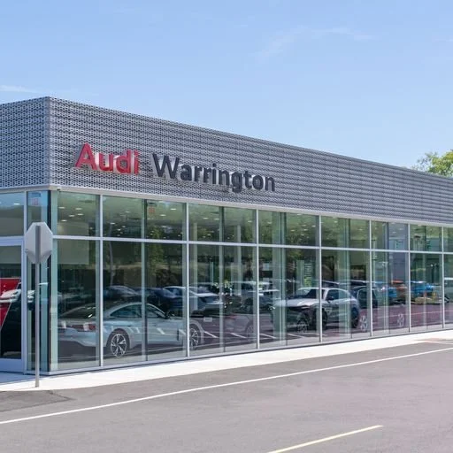 Exterior of an Audi dealership named Audi Warrington with large glass windows showing cars inside and outside.