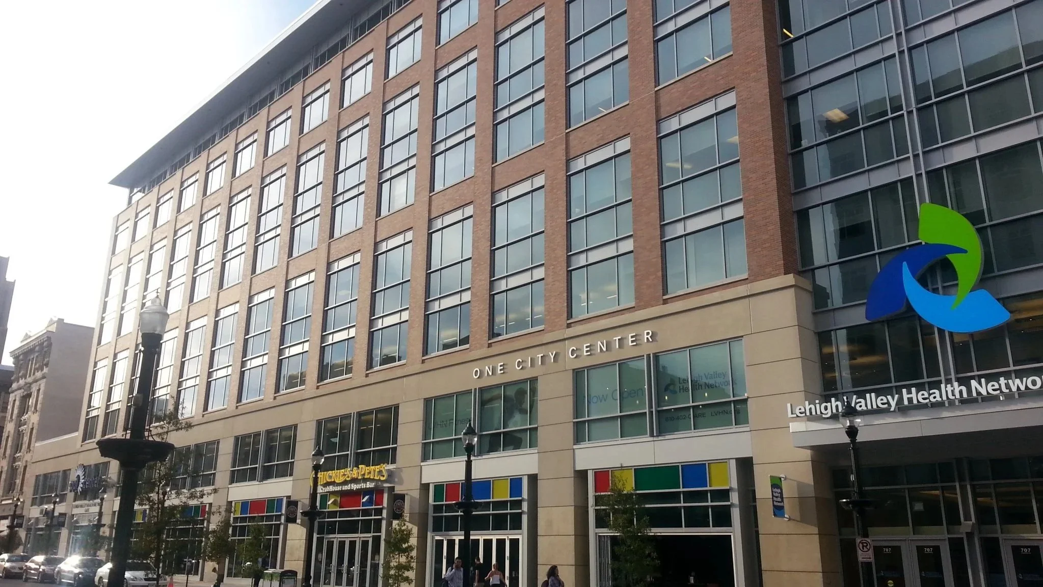 Tall modern building with glass windows and a sign that reads 'ONE CITY CENTER.' There are street lamps and pedestrians in front, with a colorful logo of green and blue in the lower right corner.