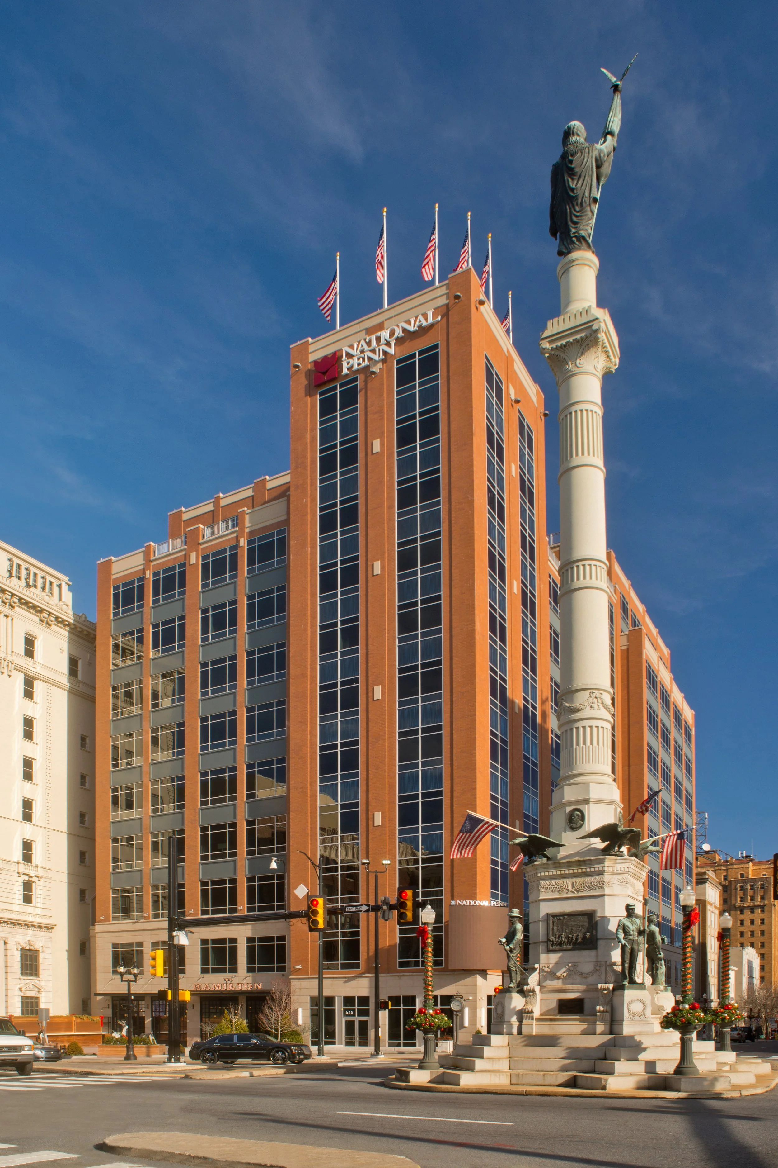 A city street corner with a monument featuring statues and an obelisk, a tall modern brick building with flags on top, and a traffic light.