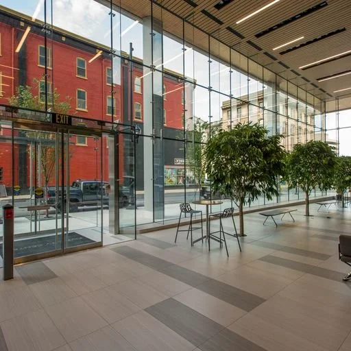 An indoor lobby or waiting area with large floor-to-ceiling glass windows, potted plants, and outdoor city street view.
