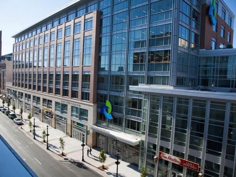 Modern multi-story office building with glass windows and commercial storefronts on the ground floor, located on a city street with parked cars and sidewalk trees.