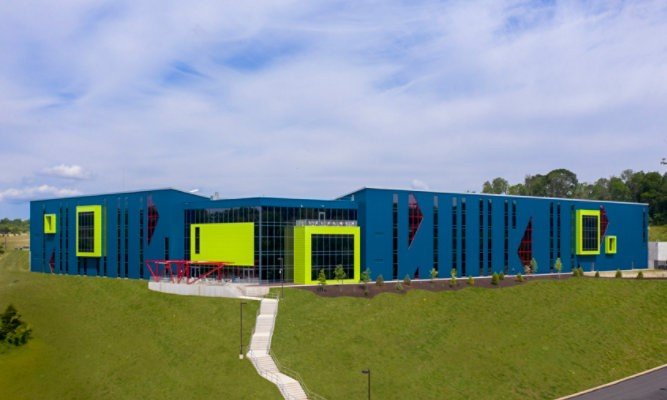 Modern blue school building with large green accents, windows, and a sloped walking path leading to the entrance, situated on a grassy hill under a partly cloudy sky.