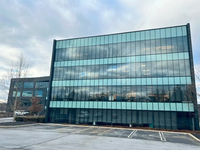 A modern multi-story office building with glass windows, reflecting the cloudy sky and trees, and a parking lot in the foreground.