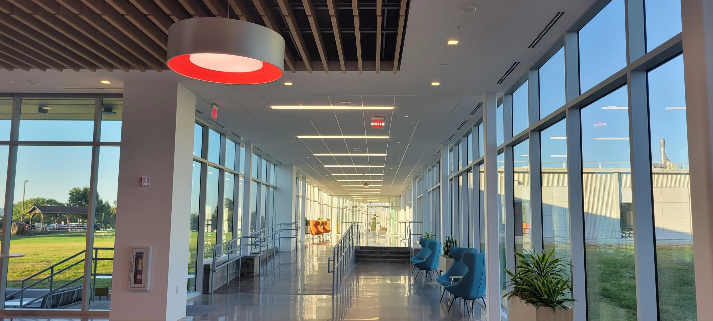 Bright, modern indoor hallway with large floor-to-ceiling windows on both sides, blue chairs, and plants, illuminated by natural sunlight and ceiling lights.