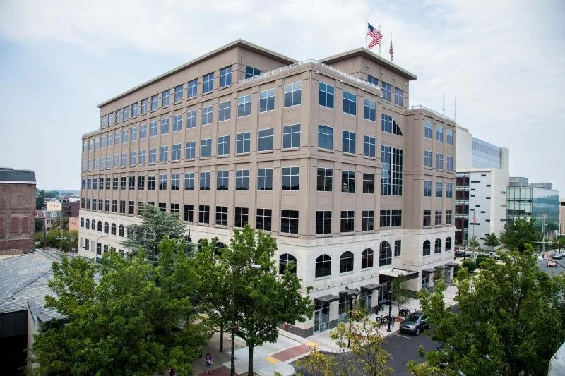 Multi-story office building with American flags on top, surrounded by trees and a street with cars.