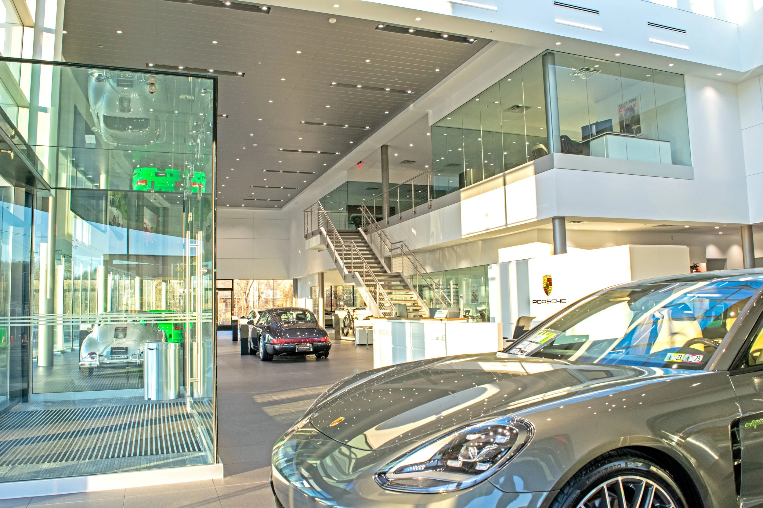 Interior of a modern Porsche dealership showroom with luxury cars on display, including a silver Porsche in the foreground and other cars on the showroom floor. There are glass walls, a staircase, and a second-floor balcony with offices or display ar